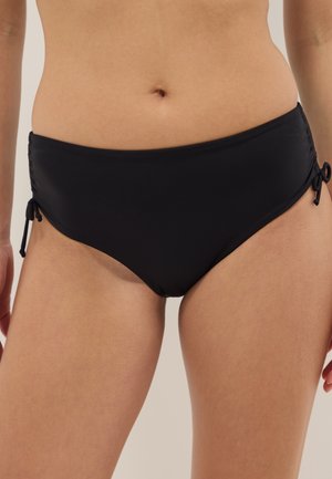 Vrouw draagt zwarte bikinibroekjes met zijstrikdetails, toont het middel en de bovenbenen tegen een neutrale achtergrond.