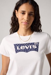 Balta medvilninė marškinė su tamsiai mėlynomis ir baltomis taškeliais Levi's logotipu. Išvaizdą puikiai papildo sluoksniuotos auksinės grandinėlės.