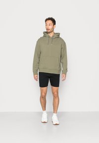 Olivgrüner Pullover mit Kapuze und Fronttasche, kombiniert mit schwarzen Shorts und weißen Sneakers. Glattes Material und lockere Passform.