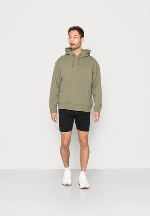 Olivgrüner Pullover mit Kapuze und Fronttasche, kombiniert mit schwarzen Shorts und weißen Sneakers. Glattes Material und lockere Passform.