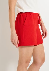 Shorts rouges texturés avec une taille élastique, dotés de poches latérales et d'un détail de pompon sur la taille.