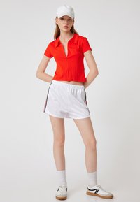 Rode cropped polo shirt met een ritssluiting, gecombineerd met witte sportshorts met zwarte en rode zijstrepen. Witte pet en sneakers.