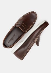 Brown leather moccasin-style shoes with white rubber soles, decorative stitching, side cutouts, and gold-tone eyelets.
