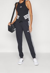 Černý atletický nátělník s bílými pruhy, doplněný tmavými tepláky se stranovými pruhy. Doplňky zahrnují černou sportovní tašku a bílé tenisky.