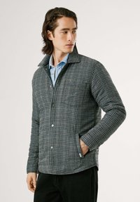 Joven con cabello largo y castaño, vistiendo una chaqueta de cuadros y una camisa de denim, posa con confianza frente a un fondo neutro.
