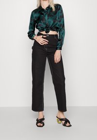 Grünes, blumenmusteriges Satinhemd, am Bund geknotet; schwarz-hochgeschnittene Jeans mit weißen Nähten; schwarze Sandalen mit verflochtenen Riemen und goldenen Akzenten.