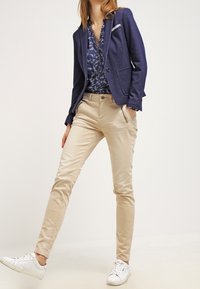 Granatowa marynarka z dwoma przednimi kieszeniami, wzorzysta bluzka, beżowe, dopasowane spodnie oraz białe tenisówki, prezentujące proste linie i gładkie tekstury.