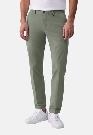 Uomo che indossa chino verde oliva chiaro con orli risvoltati e sneakers bianche, in piedi davanti a uno sfondo chiaro e uniforme.