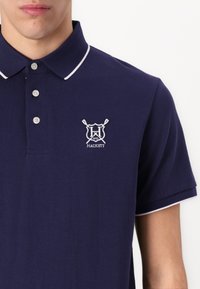 Tamsiai mėlynas polo marškinėliai su tekstūriniu paviršiumi, balta apykaklės danga, trims mygtukais ir logotipu, kuriame pavaizduoti kryžiuoti irai ir prekės ženklas "Hackett."