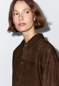 A woman with shoulder-length brown hair gazes slightly to the side, wearing a dark brown shirt against a plain white background.