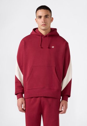 Junger Mann, der einen roten Hoodie mit weißen schrägen Streifen auf den Ärmeln und passende rote Hose trägt, steht vor einem schlichten weißen Hintergrund.