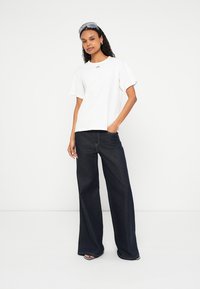 Άσπρο βαμβακερό t-shirt με κοντά μανίκια και μικρό λογότυπο, συνδυασμένο με σκούρα τζιν παντελόνια φαρδιάς γραμμής. Μινιμαλιστικός σχεδιασμός και άνετη εφαρμογή.