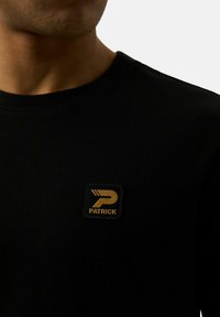 Czarny bawełniany t-shirt z okrągłym dekoltem, z kwadratową złotą łatą z napisem „PATRICK” oraz stylizowanym logo na lewej piersi. Miękka tekstura.
