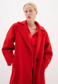 Cappotto rosso in lana con ampio colletto a risvolto, chiusura con un bottone e vestibilità leggermente ampia. Indossato sopra una blusa rossa, presenta tasche laterali.