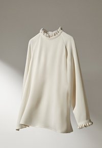 Top crème à manches longues avec un col et des poignets volantés. Fabriqué dans un tissu doux et lisse, il offre une coupe décontractée et un ourlet légèrement arrondi.