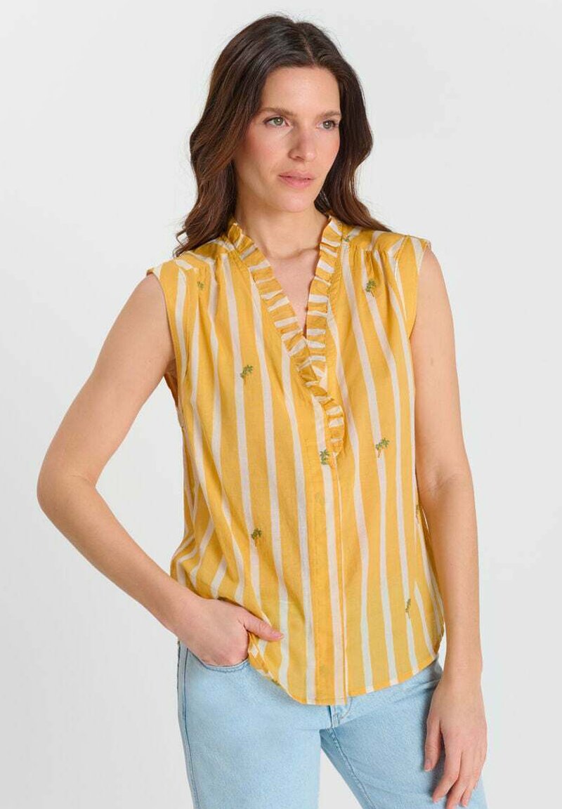 Femme portant un chemisier jaune sans manches à rayures verticales blanches et petits imprimés de palmiers verts, associé à un jean bleu clair.