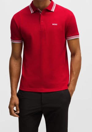 Man draagt een rood poloshirt met korte mouwen met een witte gestreepte kraag en mouwboorden, en een klein "BOSS"-logo op de borst, gecombineerd met een zwarte broek.