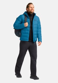Blaues Puffy-Jackett mit Reißverschluss und Kapuze, getragen über einer schwarzen Basisschicht. Kombiniert mit schwarzen Hosen und schwarzen Wanderschuhen.