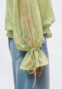 Διάφανη πράσινη μπλούζα με floral μοτίβο, μανίκι με ελαστική μανσέτα και δεμένο φιόγκο, φορεμένη με ανοιχτόχρωμο τζιν παντελόνι.