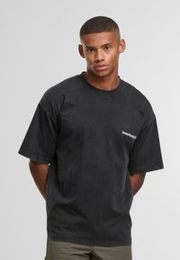 Černé oversized tričko vyrobené z bavlny s krátkými rukávy, s malým bílým logem na levé hrudi. Měkká textura a ležérní design.