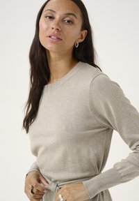 Pull beige à manches longues, col rond, avec un détail à nouer à la taille. Texture lisse avec des côtes subtiles le long des poignets.