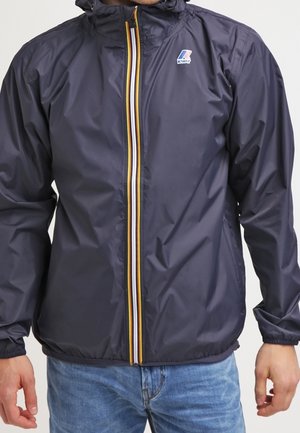 Granatowa kurtka wiatrówka z przednim zamkiem błyskawicznym, ozdobiona żółtymi, białymi i niebieskimi paskami, noszona na jasnoniebieskich dżinsach.