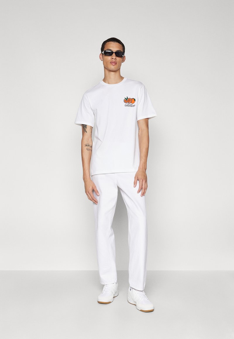 Camiseta blanca de algodón con un diseño gráfico de naranjas, combinada con pantalones blancos y zapatillas blancas. El modelo lleva gafas de sol oscuras.