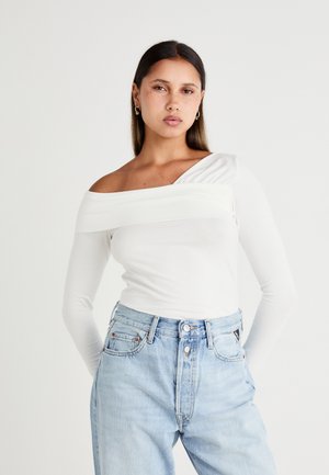 Witte long sleeve top met een off-shoulder design, voorzien van een geplooid accent op één schouder. Gecombineerd met lichtblauwe high-waisted jeans.