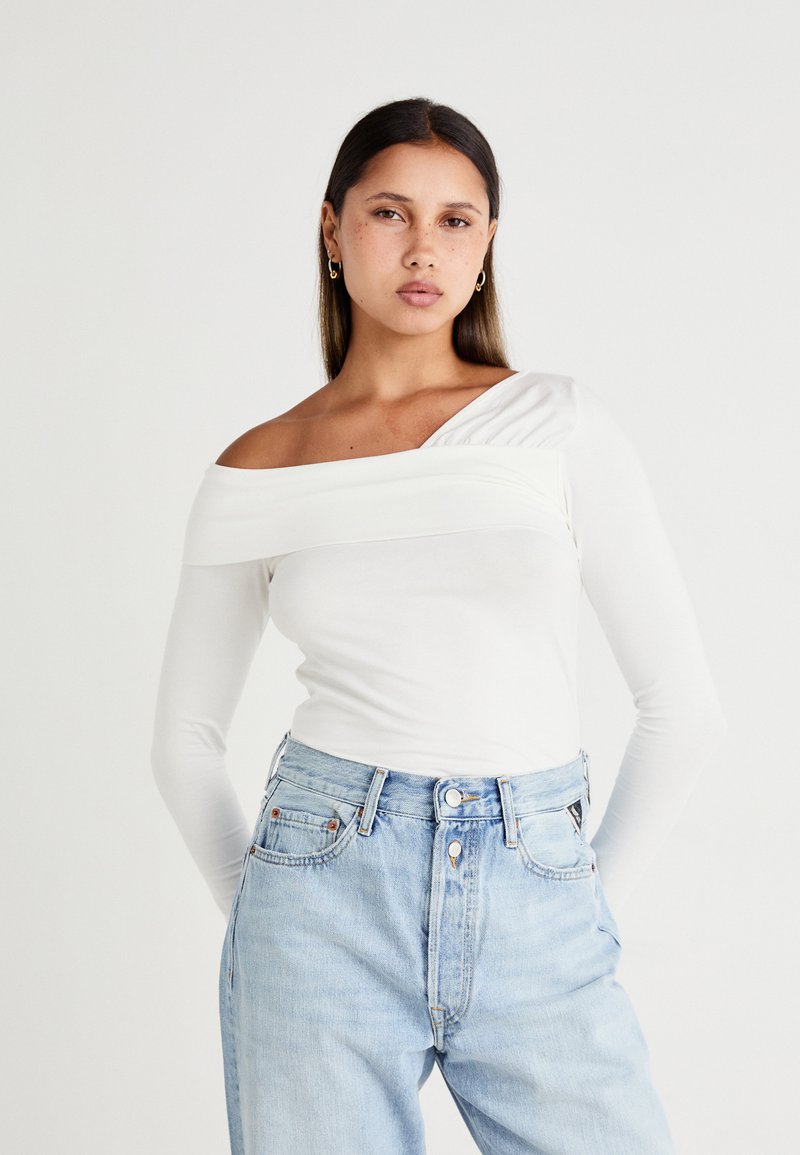 Witte long sleeve top met een off-shoulder design, voorzien van een geplooid accent op één schouder. Gecombineerd met lichtblauwe high-waisted jeans.