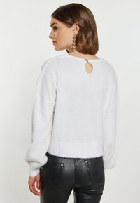 Valkoinen, pörröinen neule, jossa on pyöreä takakainaloketju ja tiukka ribbibandi, yhdistettynä kiiltäviin mustiin slim fit -housuihin.
