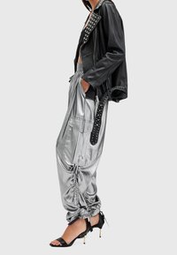 Personne portant un pantalon cargo argent froncé, une veste en cuir noire cloutée et des sandales noires à talons hauts avec brides à la cheville, debout de profil.