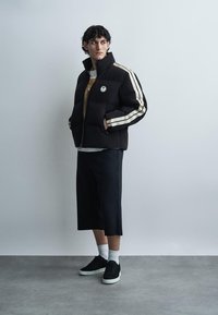 Junger Mann trägt eine schwarze Steppjacke mit weißen Streifen, schwarze weite Shorts, weiße Socken und schwarze Turnschuhe, steht vor einer schlichten Wand.