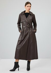 Bruine leren trenchcoat met een dubbelbreasted ontwerp, brede revers, een ceintuur om de taille en zijzakken. Zwarte enkellaarsjes complementeren de outfit.