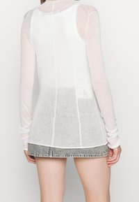 Lightweight long-sleeve top in sheer white fabric, featuring a fitted design, contrasting gray hem, and a subtle logo detail at the back.
