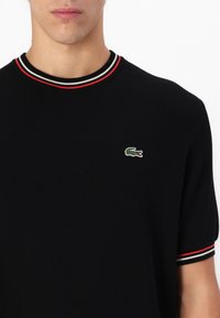 Black knitted short-sleeve t-shirt with a ribbed collar featuring red and white stripes. Includes a small green embroidered crocodile logo.