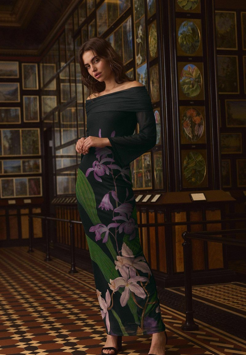 Sort off-the-shoulder kjole med blomsterdesign i lilla og grøn, med en tætsiddende silhuet og semi-gennemsigtige lange ærmer.