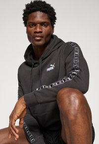 Zwarte hoodie gemaakt van zachte stof, met een kangoeroezak, capuchon met trekkoorden en een contrasterend wit Puma-logo en zijtapedesign.