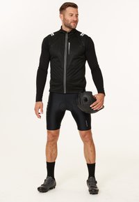 Svart cykelväst med reflexaccenter, bärs över en långärmad topp, kombineras med svarta shorts och cykelskor, håller en hjälm.