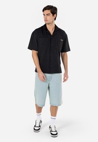 Camisetta nera a maniche corte con due tasche frontali, tessuto testurizzato e chiusura a bottone, abbinata a pantaloni blu chiaro in denim e sneakers nere.