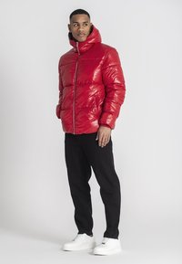 Rote Pufferjacke mit glänzender Oberfläche, hohem Kragen, Frontreißverschluss und Taschen. Kombiniert mit schwarzen Hosen und weißen Turnschuhen.