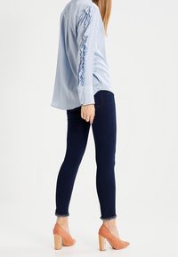 Blouse bleu clair à manches longues avec des détails volantés sur les manches, associée à un jean skinny foncé et des talons de couleur pêche avec un design de talon bloc.