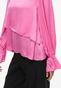 Rosa lagdelt bluse i lett, teksturert stoff med lange, puffede ermer og rynkede mansjetter, båret over svarte bukser.