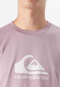 Egy személy nyaka és felső melle látható, lila színű pólót visel, amelyen egy nagy fehér Quiksilver logó található.