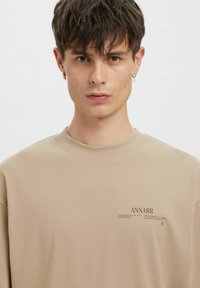 Béžová bavlněná mikina s kulatým výstřihem, volným střihem a tištěným textem na levé straně, s jemnou texturou po celé ploše.