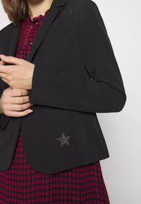 Černý blazer s třpytivými hvězdnými detaily na ramenech a dolním lemu, nošený přes šaty s červeným a černým vzorem. Hladká textura.