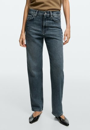Femme portant un jean gris foncé taille haute à jambes droites, un top marron et des ballerines noires pointues, debout devant un fond uni.