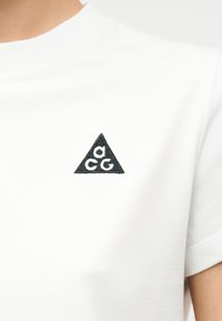 White crewneck t-shirt with a small black triangle emblem featuring three lowercase letters "a," "c," and "g" on the left chest.