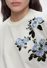 Gros plan d'une personne portant un haut écru avec une broderie florale bleue détaillée et des feuilles vert foncé sur la zone des épaules.