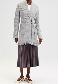 Šedý žebrovaný pletený kardigan s páskem na zavazování, otevřeným předním dílem a třásněmi na spodním okraji. Kombinován s plisovanou tmavě šedou sukní a světlými balerínkami.