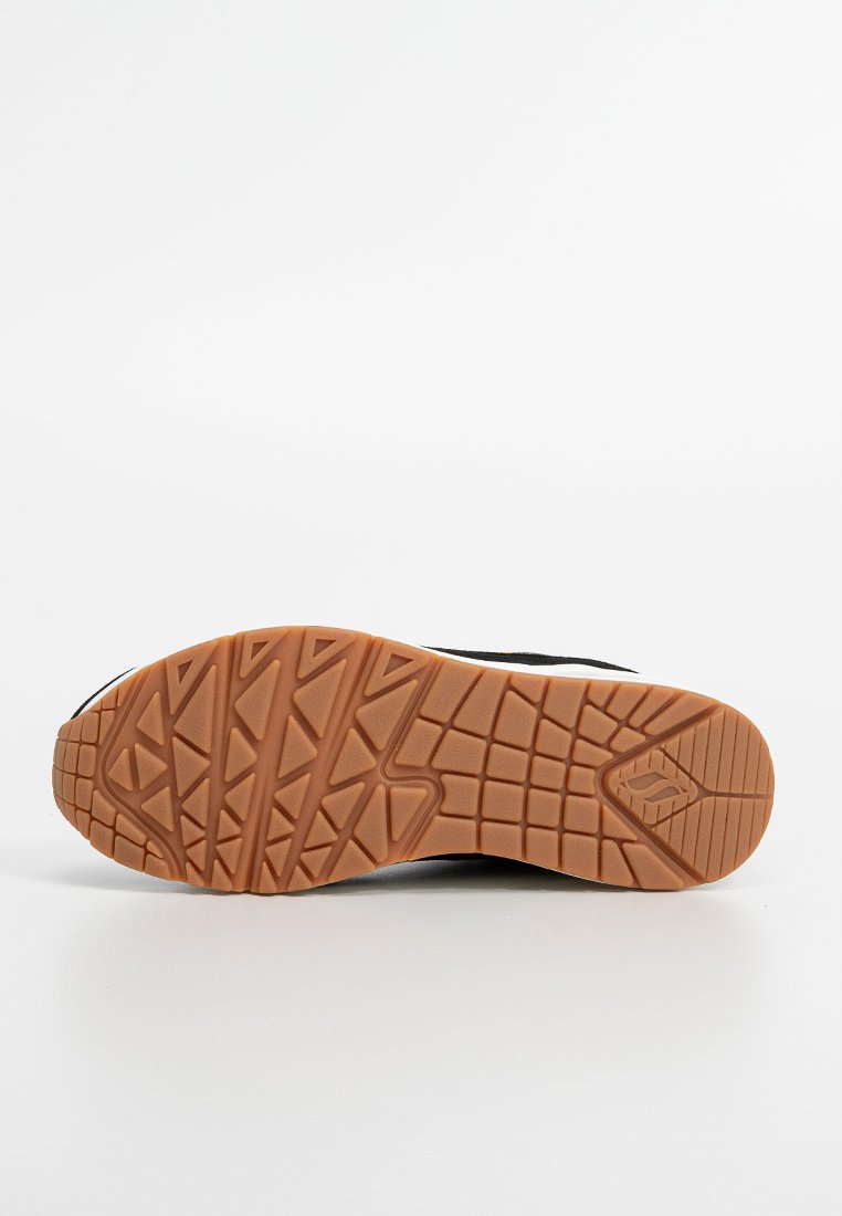 Bruine rubberen zool van een zwarte schoen met geometrische profielpatronen en een klein logo nabij de teen.